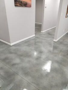 A hallway with polished concrete floors reflecting the overhead lights. The walls are painted light grey, and there's a framed painting hanging on the left wall. The hallway has several doorways leading to adjacent rooms.