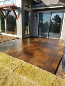 A modern house features large glass doors leading to an outdoor patio with a glossy, stained concrete floor in shades of brown. The patio adjoins a green lawn, and the house exterior is gray with wood and metal accents.