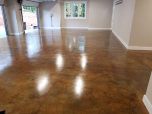 A newly finished empty garage with polished concrete floors. The space is well-lit by natural light coming through a window and open garage door. The walls are painted light beige, and the garage has a clean and spacious appearance.