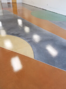 A polished concrete floor featuring an abstract design with various geometric shapes and colors, including blue, yellow, green, and orange. The surface has a glossy finish, reflecting the overhead lights.