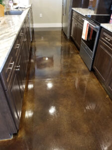 A modern kitchen with polished concrete floors, dark wooden cabinets, and marble countertops. The floor is highly reflective, showing off a glossy, smooth surface. Stainless steel appliances, including a refrigerator and oven, are visible on the right side.